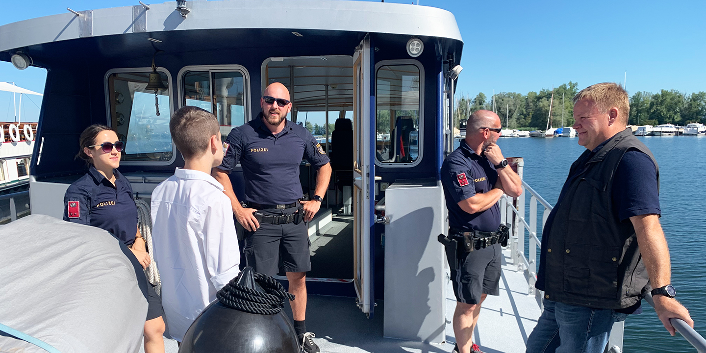 Teo steht mit Gert Gröchenig von der Landespolizeidirektion Vorarlberg und anderen Polizisten auf dem Deck eine Polizeibootes.