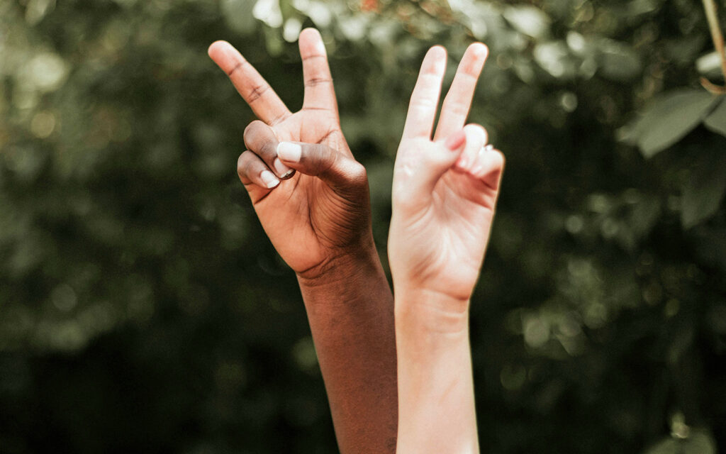 Zwei H&auml;nde in zwei unterschiedlichen Hautt&ouml;nen strecken in die H&ouml;he und zeigen das Peace-Zeichen