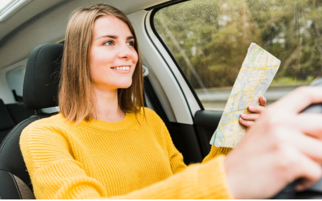 Eien junge Frau in einem gelben Pullover sitzt am Steuer und h&auml;lt eine Landkarte in der Hand.