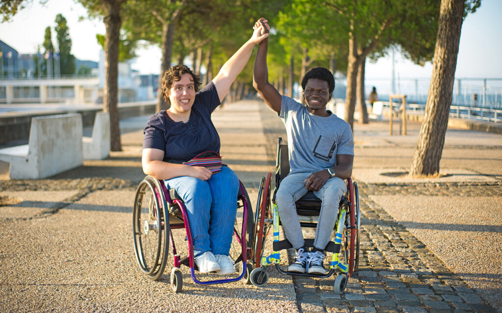 Ein Junge und ein M&auml;dchen sitzen im Rollstuhl an der Seepromenade und geben sich HIgh 5