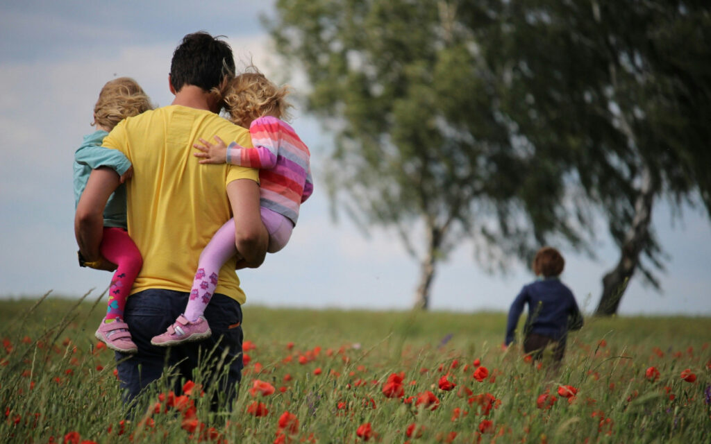 Ein Vater steht mit zwei Kindern im Arm in einem Mohnfeld und vor ihm rennt ein weiteres Kind.